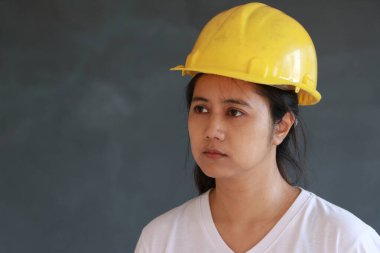 Kadın inşaat mühendisi ya da mimar sarı kasklı ve koyu gri arka planda beyaz tişört giyiyor.. 