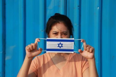 A woman with Israel flag on hygienic mask in her hand and lifted up the front face on blue background. Tiny Particle or virus corona or Covid 19 protection. Concept of Combating illness. clipart