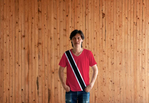 Man wearing Trinidad and Tobago flag color shirt and standing with two hands in pant pockets on the wooden wall background, a red field with a white-edged black diagonal band.
