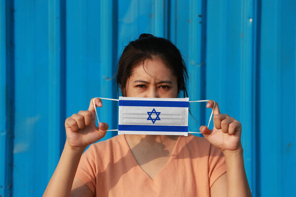 A woman with Israel flag on hygienic mask in her hand and lifted up the front face on blue background. Tiny Particle or virus corona or Covid 19 protection. Concept of Combating illness.