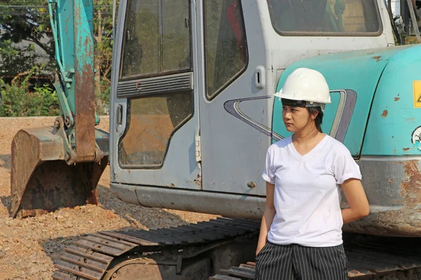 女性土木工程师或建筑师身穿白色头盔和白色T恤 站在工作区的绿色挖掘机车旁边 — 图库照片