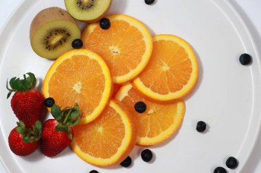 İnce yuvarlak portakal dilimleri beyaz tabakta çilek, kivi meyvesi ve yaban mersiniyle bir araya toplanır..