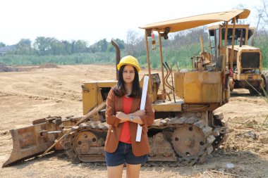 Kadın inşaat mühendisi ya da mimar, iş yerindeki ön yükleme mühendisliği inşaat aracının arka planında dururken sarı miğferi takar ve proje taslaklarıyla kollarını çaprazlar..