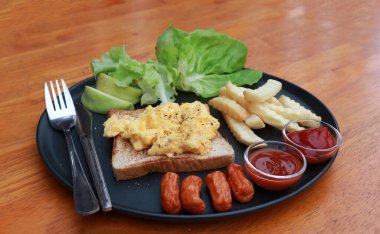 Tahta zemindeki siyah yuvarlak tabakta kahvaltı. Yağda yumurta ve sosisli ekmek, ketçaplı patates kızartması ve acı sos, marul ve guava..