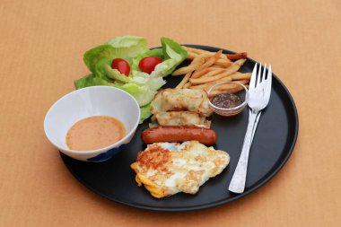 Kahverengi zemindeki siyah yuvarlak tabakta kahvaltı. Sosisli sahanda yumurta, patates kızartmalı mantı ve domatesli sebze..