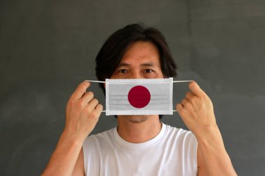 Elinde Japon bayrağı olan hijyenik maskeli bir adam koyu gri arka planda ön yüzünü kaldırdı. Minik parçacık ya da virüs koronası ya da Covid 19 koruması. Hastalıkla mücadele kavramı.