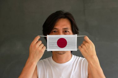 A man with Japan flag on hygienic mask in his hand and lifted up the front face on dark grey background. Tiny Particle or virus corona or Covid 19 protection. Concept of Combating illness. clipart
