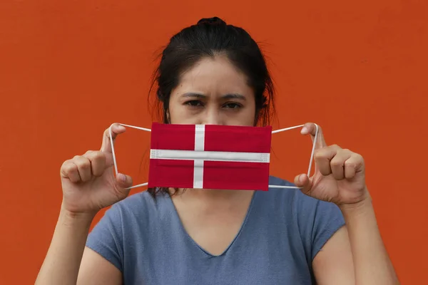 一名妇女手里拿着丹麦国旗 戴着卫生面具 用橙色的背景把前面的脸抬起来 微小颗粒或病毒电晕或Covid 19保护 防治疾病的概念 — 图库照片