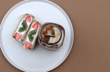 Kahverengi zeminde yuvarlak tabağın beyaz renginde kahvaltı. Çilek ve kremalı sandviç, bir bardak hindistan cevizli buzlu kahve..