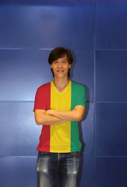 Man wearing Guinea flag pattern shirt and cross one's arm on the blue wall background. A vertical tricolor of red yellow and green.