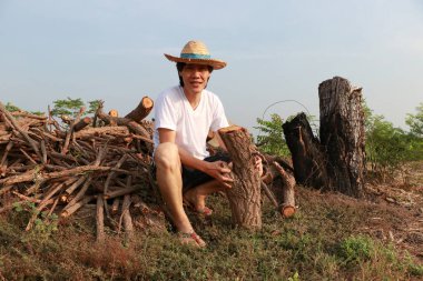 Şeker kamışı çiftliğindeki odun yığınında kerestelerle oturan ve beyaz tişörtlü hasır şapka giyen bir çiftçi..