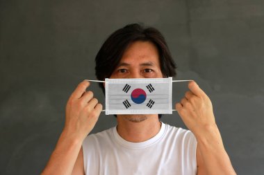 A man with South Korea flag on hygienic mask in his hand and lifted up the front face on dark grey background. Tiny Particle or virus corona or Covid 19 protection. Concept of Combating illness. clipart