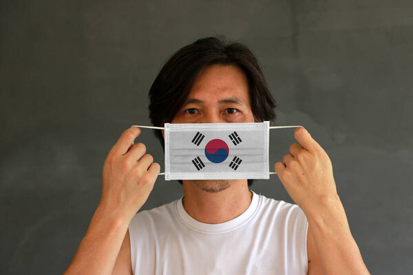 A man with South Korea flag on hygienic mask in his hand and lifted up the front face on dark grey background. Tiny Particle or virus corona or Covid 19 protection. Concept of Combating illness.