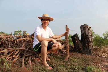 Şeker kamışı çiftliğindeki odun yığınında kerestelerle oturan ve beyaz tişörtlü hasır şapka giyen bir çiftçi..