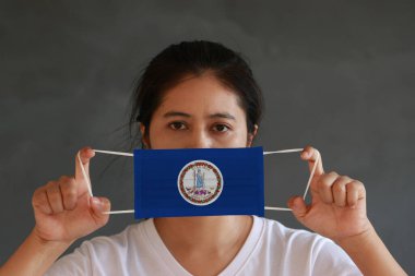 Elinde Virginia bayrağı deseni olan bir kadın ve hijyenik maske koyu gri arka planda yüzünü kapatmak için kaldırıyor. Maske, Tiny Partikül veya virüs koronası veya Covid 19 'a karşı çok iyi bir korumadır..