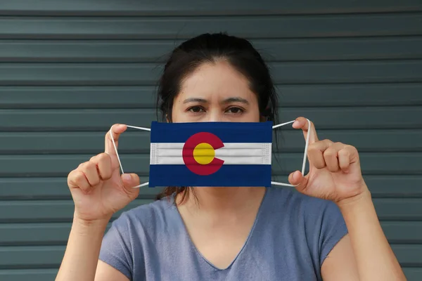 A woman and hygienic mask with Colorado flag pattern in her hand and raises it to cover her face on dark green background. A mask is a very good protection from Tiny Particle or virus corona or Covid 19.
