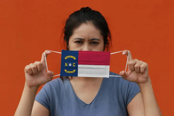 A woman and hygienic mask with North Carolina flag pattern in her hand and raises it to cover her face on orange color background. A mask is a very good protection from Tiny Particle or virus corona or Covid 19.