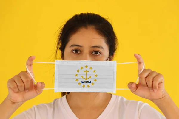 A woman and hygienic mask with Rhode Island flag pattern in her hand and raises it to cover her face on yellow background. A mask is a very good protection from Tiny Particle or virus corona or Covid 19.