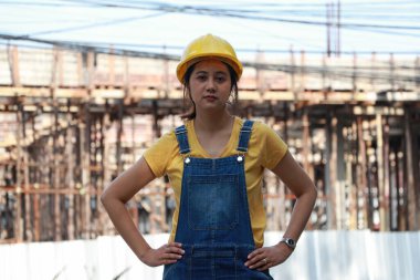 İnşaat mühendisi ya da mimar sarı kasklı, ayakta duran ve çalışma alanında akimbo yapan bir kadın..