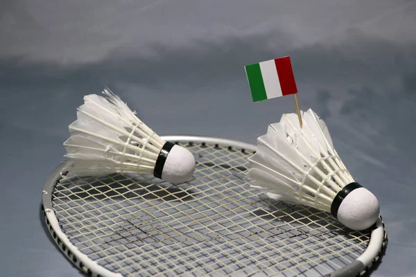 Mini Italy flag stick on the shuttlecock and another shuttlecock put on the net of badminton racket on grey floor.