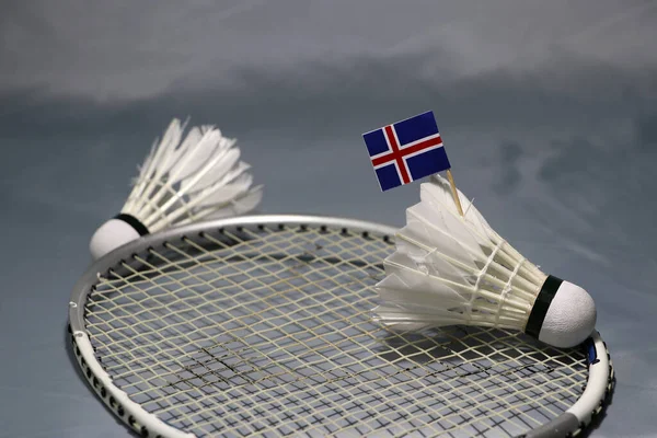 Mini Iceland flag stick on the shuttlecock put on the net of badminton racket and out focus a shuttlecock on the grey floor.