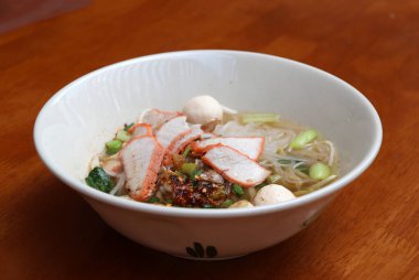 Pirinç çubuklu şehriye çorbası, ahşap masadaki kasede kırmızı domuz rostosu..