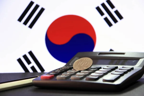 One hundred Korean won coin on reverse, (KRW) on black calculator with black and white pencil on black floor and Korean flag background. the concept of finance.