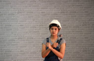 Teknisyen kadın, gri tişörtlü, kot pantolonlu, önlüklü gri tuğla desenli arka planda çelik çekiçli beyaz miğfer giyer..