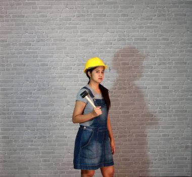Teknisyen kadın gri tişörtlü, kot pantolonlu önlüklü, gri tuğla desenli arka planda kauçuk çekiçli sarı kasklı.