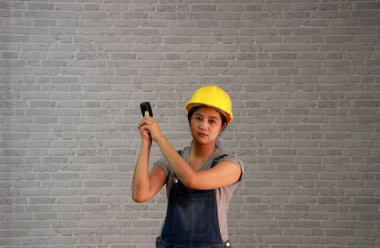 Teknisyen kadın, gri tişörtlü, kot pantolonlu, önlüklü, kauçuk çekiçli sarı miğferden iki elle gri tuğla desenli arka planda bir şeyleri parçalamayı sever..