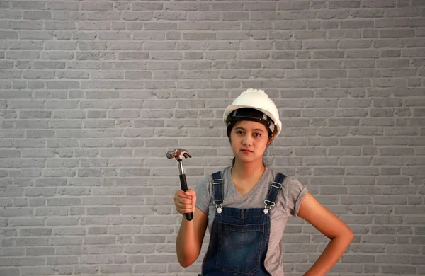 Technician woman ware white helmet with grey T-shirt and denim jeans apron dress standing and steel hammer in hand on grey brick pattern background.