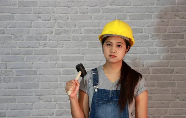 Teknisyen kadın gri tişörtlü, kot pantolonlu önlüklü, gri tuğla desenli arka planda kauçuk çekiçli sarı kasklı.