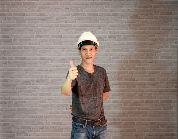 Technician man ware white helmet with dark grey T-shirt and denim jeans standing and right hand thumbs up on grey brick pattern background.