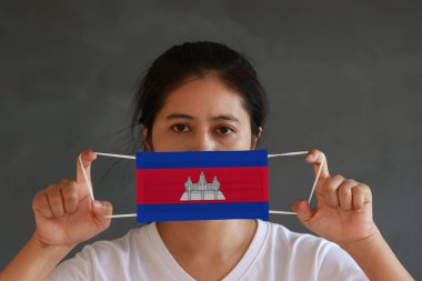 A woman in white shirt with Cambodia flag on hygienic mask in her hand and lifted up the front face on dark grey background. Tiny Particle or virus corona or Covid 19 protection. Concept of Combating illness. clipart