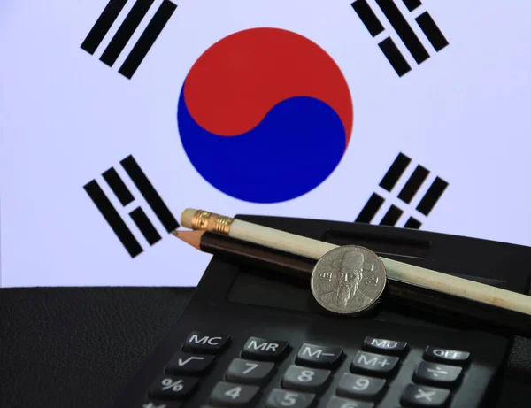 One hundred Korean won coin on obverse, (KRW) on black calculator with black and white pencil on black floor and Korean flag background. the concept of finance.