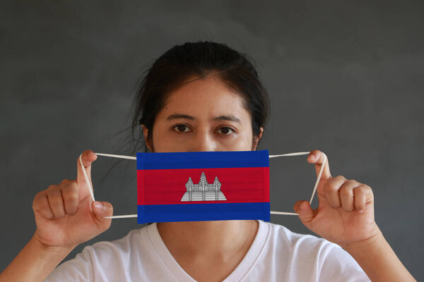 A woman in white shirt with Cambodia flag on hygienic mask in her hand and lifted up the front face on dark grey background. Tiny Particle or virus corona or Covid 19 protection. Concept of Combating illness.