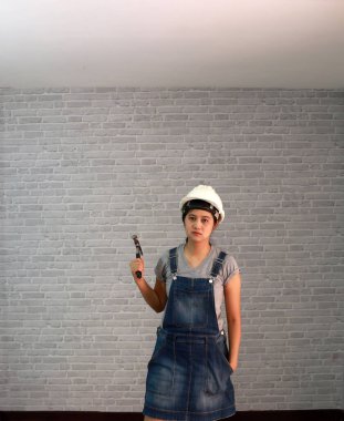Teknisyen kadın, gri tişörtlü, kot pantolonlu, önlüklü gri tuğla desenli arka planda çelik çekiçli beyaz miğfer giyer..