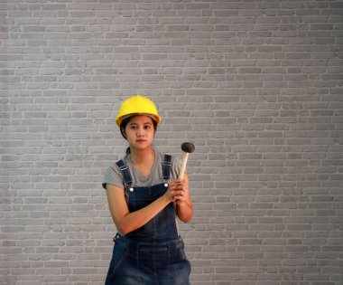 Teknisyen kadın gri tişörtlü, kot pantolonlu önlüklü, gri tuğla desenli arka planda lastik çekiçli sarı miğferli giyiyordu..