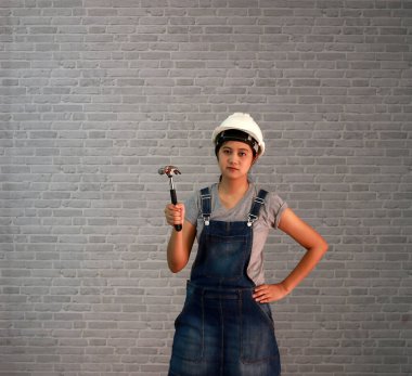 Teknisyen kadın gri tişört ve kot önlüklü beyaz miğfer gri tuğla desenli arka planda çelik çekiçli Aimbo duruyor..