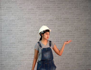 Teknisyen kadın, gri tişörtlü, kot pantolonlu, önlüklü, gri tuğla desenli arka planda avuçlarının içine bakan beyaz kasklı..