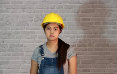 Teknisyen kadın gri tişört ve kot önlüklü sarı miğfer gri tuğla desenli arka planda duruyor..