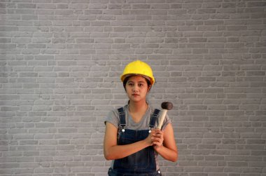 Teknisyen kadın gri tişörtlü, kot pantolonlu önlüklü, gri tuğla desenli arka planda lastik çekiçli sarı miğferli giyiyordu..