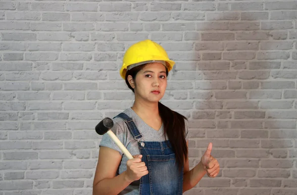 Technician woman ware yellow helmet with grey T-shirt and denim jeans apron dress standing with rubber hammer in hand and thumbs up on grey brick pattern background.