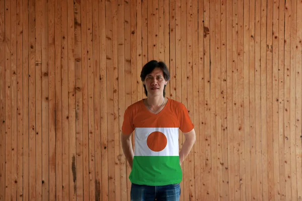 身穿尼日尔国旗色衬衫 背手交叉地站在木制墙背上的男人 中心有橙色圆圈 — 图库照片