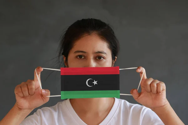 A woman in white shirt with Libya flag on hygienic mask in her hand and lifted up the front face on dark grey background. Tiny Particle or virus corona or Covid 19 protection. Concept of Combating illness.