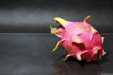 Siyah zeminde ejderha meyvesi. Şekil küreseldir. Kırmızı veya mor renkte bulunabilir. Meyvenin etrafına yeşil karanfiller yapışmış..