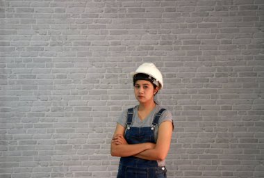 Teknisyen kadın, gri tişörtlü ve kot önlüklü beyaz kasklı 45 derecelik açıda duran ve gri tuğla desenli arka planda göğsüne sarılan bir kadın giysisi giyiyor..