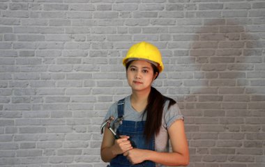 Teknisyen kadın gri tişörtlü, kot pantolonlu önlüklü, gri tuğla desenli arka planda çelik çekiçli sarı miğferli giyiyordu..