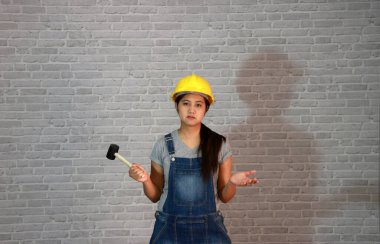 Teknisyen kadın, gri tişörtlü, kot pantolonlu önlüklü, sağ elinde kauçuk çekiç, sol elinde gri tuğla desenli arka planda avuçlar olan sarı miğfer takıyor..