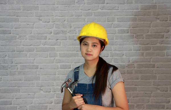 Teknisyen kadın gri tişörtlü, kot pantolonlu önlüklü, gri tuğla desenli arka planda çelik çekiçli sarı miğferli giyiyordu..